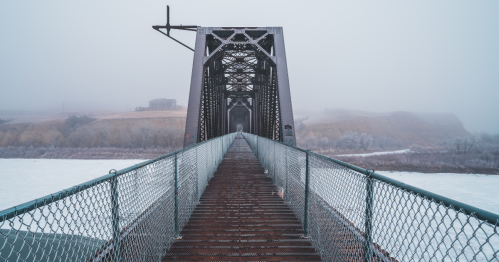 A long, abandoned railway bridge extends into a foggy, winter landscape over a frozen river.