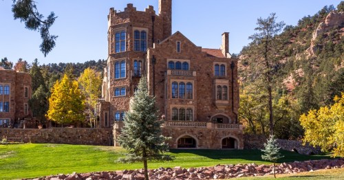Back view of Glen Eyrie Castle in Colorado Springs with tall stone towers, arched windows, and vibrant autumn trees against mountain scenery.