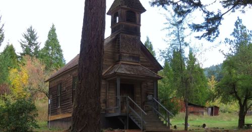 A dark wood paneled church with a bell tower in the shadows of a forest in early fall.