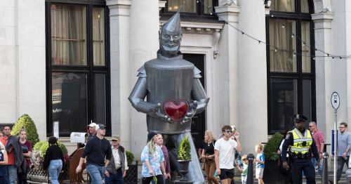 A statue of a tin man holding a red heart, and people walking past it.