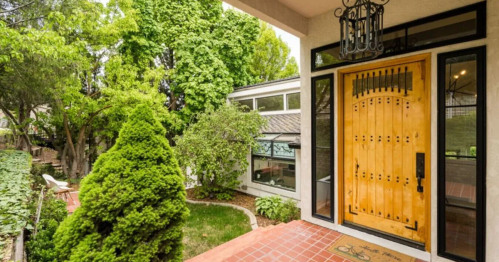Front exterior of a vacation rental in Boise, Idaho, complete with a large wooden door, iron lighting, large porch, and lush trees.