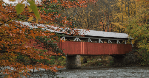 A red covered bridge spans a river framed by fall foliage in PA.