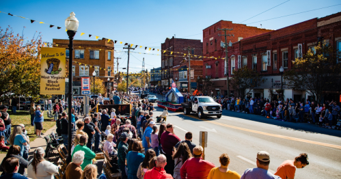 A lively parade scene with crowds lining the street, colorful flags overhead, and a tractor passing by.