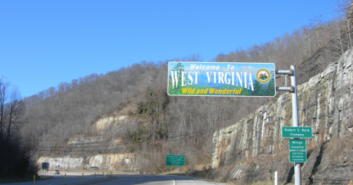 Mingo County welcome sign reads "Welcome to West Virginia" with mountain peak in the distance in the middle of winter under a blue sky.
