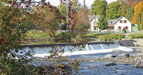 A small waterfall and river with a house on the bank, surrounded by trees with autumn leaves.