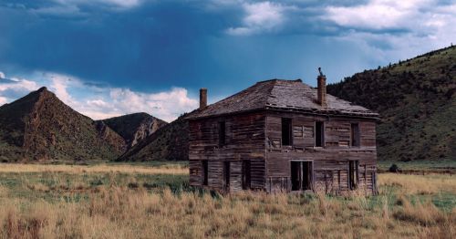 A weathered abandoned wood building in Montana, set against low foothills on dry grass prairie.