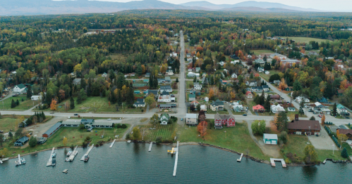 An aerial view of a small town on a lake shore surrounded by forests with fall foliage.