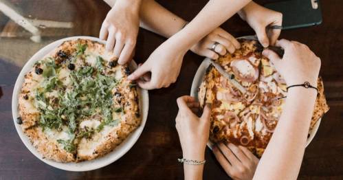 Looking down at two pizzas and several hands reaching to grab a slice.