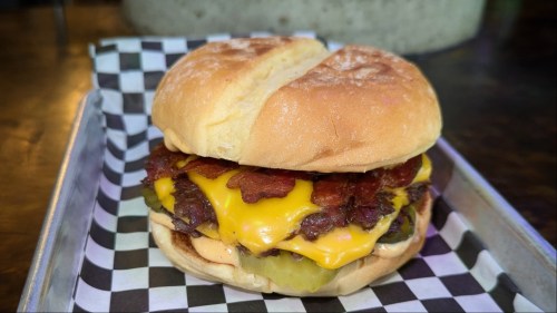 A bacon cheeseburger on black and white checkered deli paper with a toasted bun is ready to eat!
