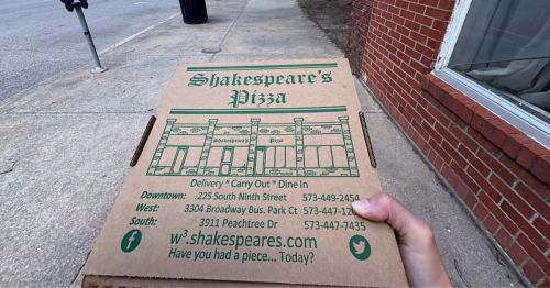 A hand holding a pizza box on a sidewalk.