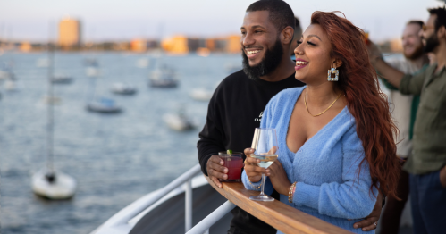 A man and woman hold drinks and gaze out at the water from an outside deck on the Spirit of Boston boat in Boston Harbor.