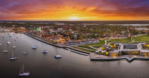 A sun setting in the distance of a small waterfront town. Boats dot the harbor. A National Monument sits to the right.