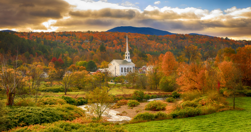 A white church steeple and a stream in Stowe, Vermont, surrounded by trees with colorful fall foliage.