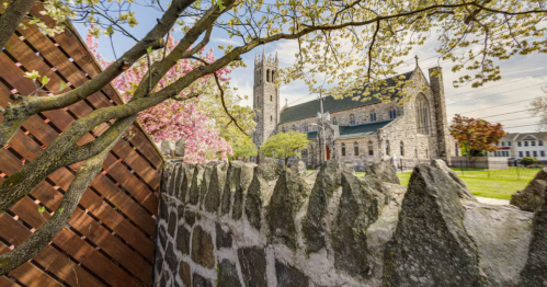 A large stone church and steeple viewed from a Vrbo patio, featuring a wooden fence and blooming trees in the foreground.