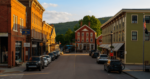 A view down a street in Waterbury, Vermont, lined with brick buildings, cars, and a distant green mountain.
