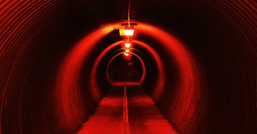 A dimly lit tunnel with curved walls is illuminated by eerie red-orange lights.