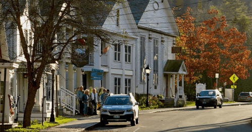 A sunny street in Wilmington, Vermont, with people walking on the sidewalk and a white building with an orange tree.
