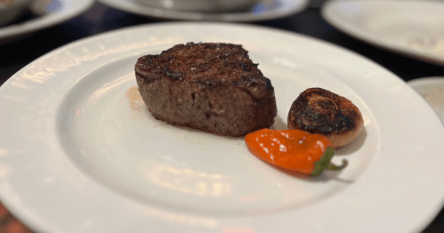 A petite steak on a white plate with a red pepper and charred garlic clove