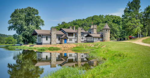 A wide shot of a medieval-style building with stone walls and a timber-framed porch, surrounded by lush green trees.