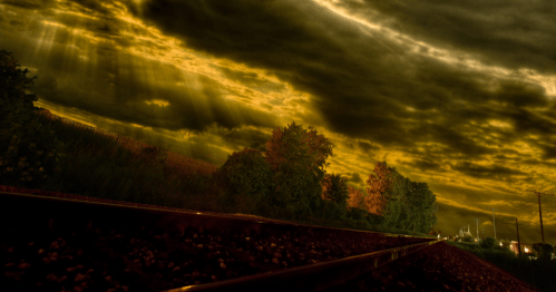 A low-angle view of two train tracks under a dramatic, dark sky with sun rays breaking through golden-hued clouds.