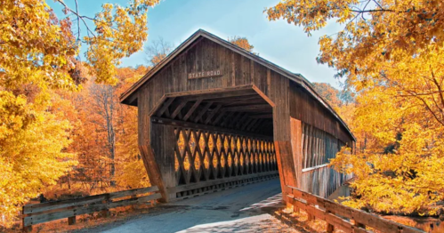 A wooden covered bridge surrounded by vibrant autumn foliage and a clear blue sky.