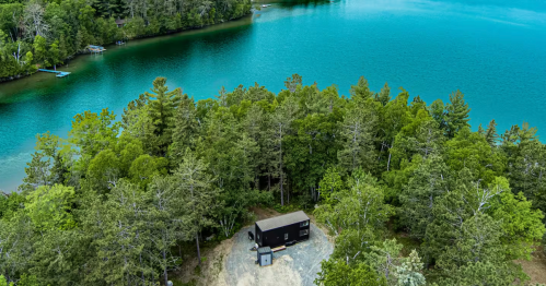 A tiny home in a clearing is surrounded by a green forest on an island in a blue lake.
