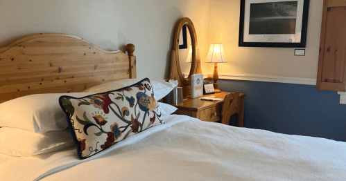 Freshly made bed with white sheets, a brown wooden headboard, and a colorful decorative pillow. The walls are white and blue with a wooden table beside the bed.