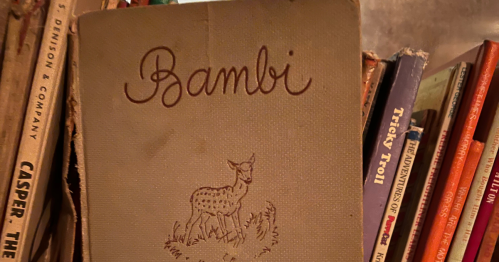 An old hardcover brown edition of Bambi with a drawing of a fawn on the front and other hardcover antique books in the background