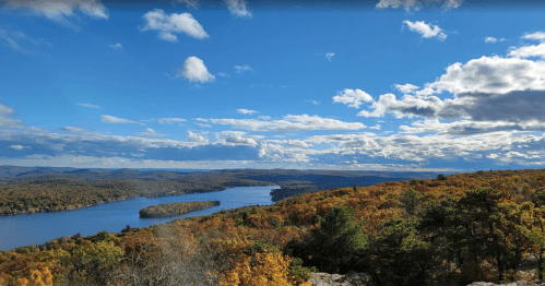 Beautiful blue lake surrounded by vibrant fall foliage under a blue sky with white clouds.