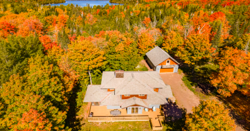 A large home and separate garage with cedar shake siding and gray roofs are surrounded by trees bursting with fall colors.