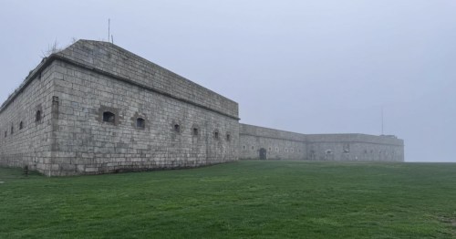 A looming, stone military base sits on a green hill covered in mist.