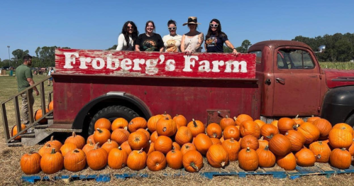 a group of 5 women stand on the bed of an old truck with a sign for froberg's farm and, in front of that truck on the ground is a pile of pumpkins