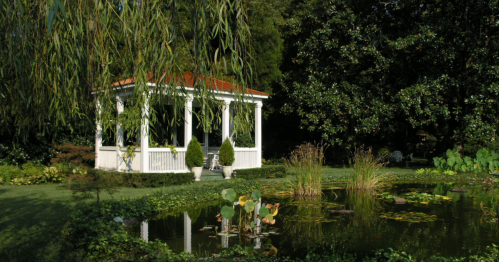 White gazebo with a red roof sits next to a small, dark pond and is partially obscured by weeping willow branches