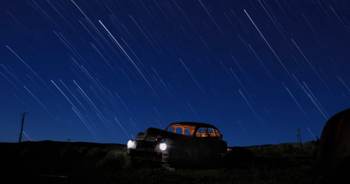 an old car with headlights and interior lights on sits in the desert under a starry sky for an astrophotography shot