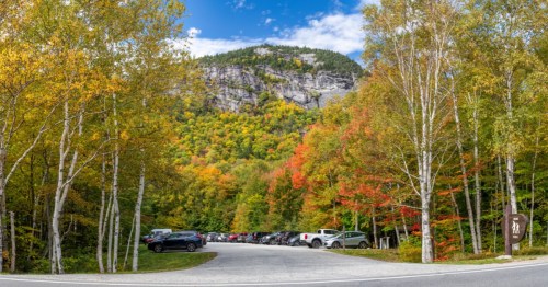 A large parking lot near a trailhead is surrounded by a forest with leaves changing into fiery colors.