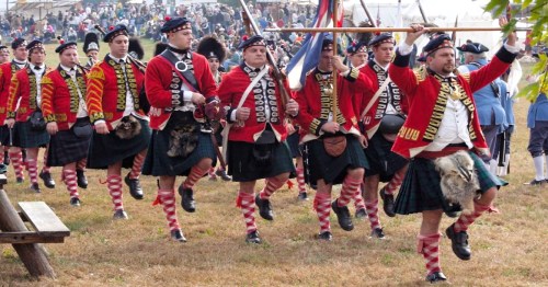 A group of soldiers in historical uniforms march in a parade, with flags and a crowd in the background.