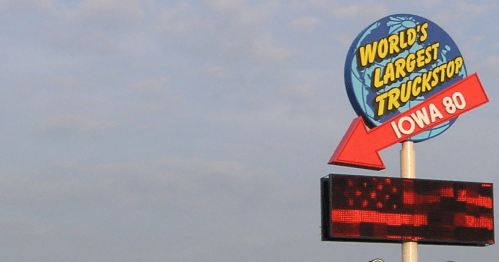 World's largest truckstop sign with a large red arrow pointing to the left towards the building against a cloudy sky.