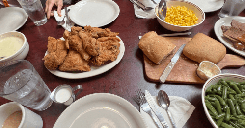 A spread of food across a wooden table. White plates surrounded by brown fried chicken, tan baked bread, and yellow corn and green beans.