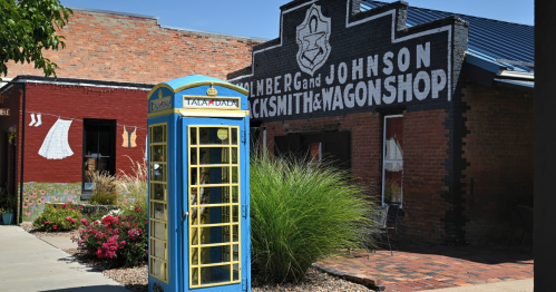 A blue and yellow old-fashioned telephone booth in the foreground with a red brick wagon shop behind it to the right and a mural of laundry drying on the line on the lefthand side.