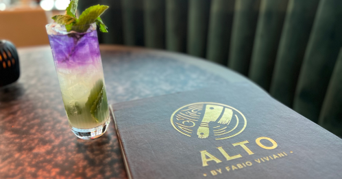 A purple and green cocktail on the left with a brown leather menu on the right. The menu reads ALTO by Fabio Viviani in gold text