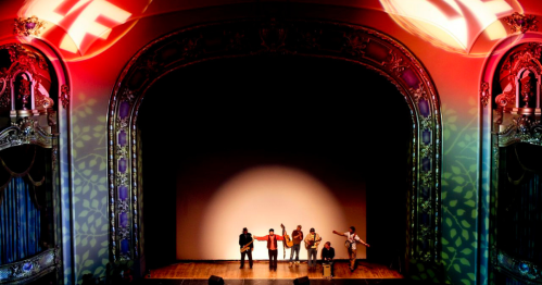 A stage lit by a spotlight with red T/F logos on either side of the stage and leaf light symbols around the bottom of the theater space