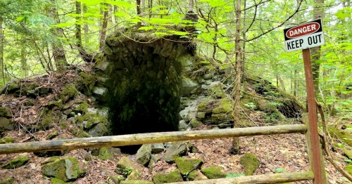 A pile of rubble lies behind a wooden rail fence at the opening of an abandoned mine shaft; a sign reading "Danger Keep Out" stands nearby.