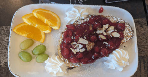 Orange slices and green grapes on the left side of a white platter with four dabs of white whipped cream around a sandwich covered in cherry preserves.