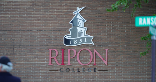 A red brick wall with a red, black, and white glossy sign that reads "1851 Ripon College"