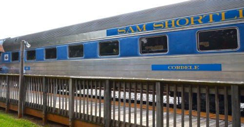 A silver and blue train car inscribed with the words "SAM Shortline" waits at a depot for passengers to board.