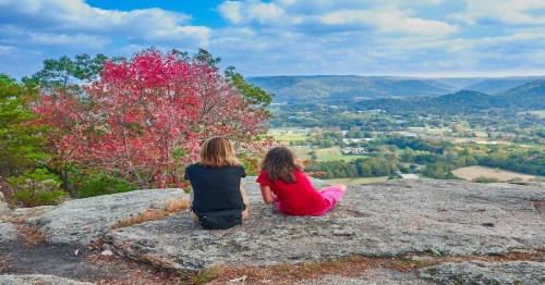 Two hikers sit on a rocky overlook and enjoy a view.
