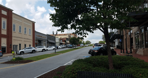A view of a historic, two-story downtown street lined with cars, storefronts, and a large shade tree.