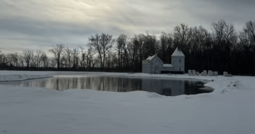 A stone castle in a winter scene, surrounded by snow and a frozen or partially frozen pond.