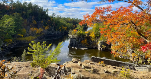 A scenic view of a river surrounded by autumn foliage and rocky cliffs under a partly cloudy sky.