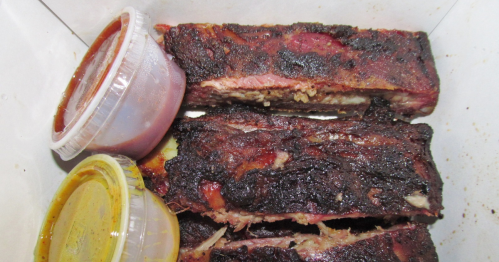 A takeout container with several pieces of St. Louis-style BBQ ribs and two small cups of sauce.